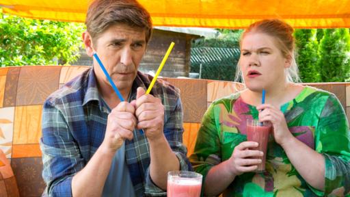 Fritz Fuchs sitzt mit einer Frau auf einer Hollywoodschaukel. Sie hält ein Saftglas mit Strohalm und er hält zwei Strohalme in der Hand und schaut die Halme nachdenklich an. 