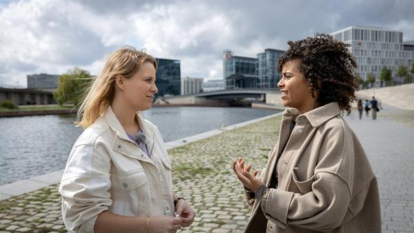 Moderatorin Linda Joe Fuhrich steht im Profil links und interviewt die Aktivistin Sarah, die rechts im Profil zu sehen ist. Hinter den beiden sind Gebäude in Berlin zu sehen.