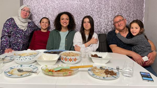 Maral sitzt mit Familie Tayan in einer Reihe an einem gedeckten Tisch. Die Teller und Schüsseln sind fast leer gegessen. Alle lächeln.