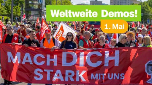 Eine Gruppe Demonstranten mit roten Fahnen und einem großen roten Banner läuft eine Straße entlang. Das große Banner trägt die Aufschrift "Mach dich stark mit uns". In der oberen Mitte des Bildes stehen die Worte "Weltweite Demos 1. Mai".