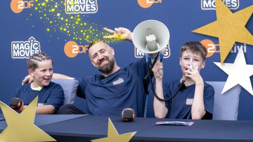 Sportreporter beim Quadball | Rechte: ZDF Gabriel (l.), Bürger Lars Dietrich (m.) und Maxi (r.) sitzen an einem Tisch. Gabriel (l.) hält ein ZDF-Mikrofon in der Hand, Maxim (r.) ein Megaphon. Im Hintergrund eine Wand auf dem das ZDF-Logo und das "Magic Moves"-
