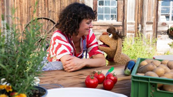 Die Landwirtin Saida vom Leckerhof und Moppi stehen zusammen hinter einem Tisch und schauen sich an. Vor ihnen auf dem Tisch befinden sich Kartoffeln, Tomaten, eine Aubergine und ein Kräutertopf. Sie befinden sich draußen, im Hintergrund ist ein Stallgebäude zu sehen.