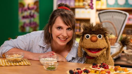 Rechte: rbb/ANDERTHALB Medienproduktion GmbH/Daria Biliak Maya und Moppi strahlen in die Kamera. Vor ihnen auf dem Tresen steht ein Brett mit "Doppimoppis". Diese Flammkuchen sind bunt belegt: auf der einen Hälfte verschiedenes Gemüse und auf der anderen Hälfte verschiedene Beeren. Auf dem Tresen befinden sich noch drei Schüsseln mit Belag-Zutaten. Im Hintergrund ist der Leckerladen mit seinen Lebensmittelregalen.