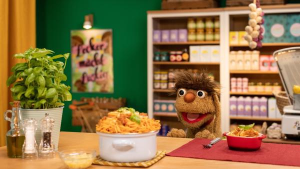 Moppi steht am Tresen im Leckerladen. Er schaut gierig auf einen großen Topf voll Spaghetti. Neben dem Topf steht unter anderem ein Basilikum, eine Flasche Öl, eine Salz- und eine Pfeffermühle. Im Hintergrund befinden sich die mit Lebensmitteln gefüllten Regale des Leckerladens.