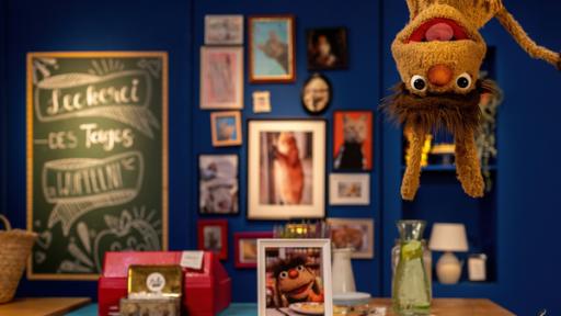 Rechte: rbb/ANDERTHALB Medienproduktion GmbH/Daria Biliak Moppis Kopf ist oben rechts im Bild zu sehen. Er scheint kopfüber von der Decke zu hängen, seine Ohren baumeln nach unten. Unter ihm steht auf dem Tresen ein Teller voller Wuffeln - herzförmige Waffeln mit einer Mangoldfüllung. Auf dem Tresen befindet sich außerdem die Kasse des Leckerladens, ein gerahmtes Bild von Moppi und eine Karaffe voller Wasser. Im Hintergrund hängen gerahmte Katzenbilder an einer Wand. An einer Tafel steht das Gericht des Tages: Wuffeln.