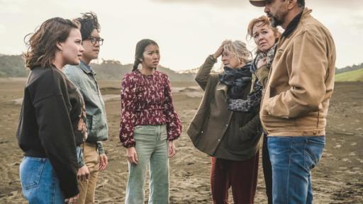 Verhängnisvolle Unterschrift | Rechte: © Libertine Pictures Limited & Slim Film + Television Limited Im sandigen Gelände stehen Issie (links), Caleb (2.v.l.) und Caroline (Mitte links) gegenüber von Mitch (Mitte rechts), Amanda (2.v.r.) und Tom (rechts). Alle schauen besorgt. Der Himmel ist grau.