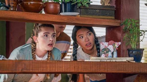Verkupplungsversuch | Rechte: © Libertine Pictures Limited & Slim Film + Television Limited Natasha (links) und Caroline (rechts) stehen hinter einem Holzregal und beobachten mit staunenden Blicken, durch die Regalbretter hindurch, was auf der anderen Seite vor sich geht.