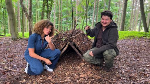Tessniem hockt gemeinsam mit Survival-Trainer Steffen vor einem selbstgebauten Unterschlupf aus Ästen und Laub.