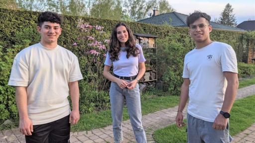 Mona steht mit Mustafa und Zubair im Garten. Sie lächeln in die Kamera.