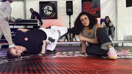 Breaking - Vom Trendsport zu Olympia | Rechte: WDR/tvision Melina Fernandez macht einen Freeze auf der Bühne der Deutschen Breaking Meisterschaft 2024 in Hamburg. Daneben sitzt Tessniem Kadiri und zeigt auf sie.