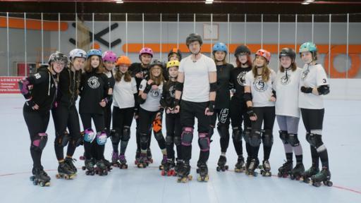 Reporter Robert und das Roller Derby Team "Mogweis Junior Roller Derby" aus Oberhausen stehen auf Rollschuhen und mit Schutzausrüstung in einer Sporthalle.