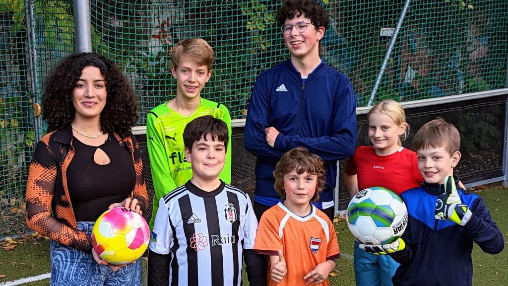 Tessniem hält einen Fußball in der Hand und steht neben einer Gruppe Kinder auf einem Fußballfeld.