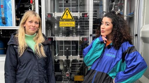 Wasserstoff - Die grüne Energie der Zukunft? | Rechte: WDR/tvision Moderatorin Tessniem steht mit der Wasserstoff-Referentin Helle Moritzen von GP Joule vor einem Elektrolyseur im Windpark in Bosbüll. Der Elektrolyseur kann Wasserstoff in die Bestandteile Wasser und Sauerstoff aufspalten.