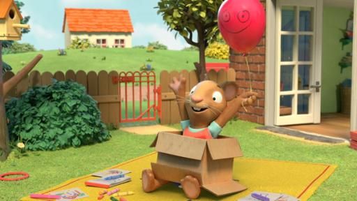 Posy sitzt freudestrahlend in einem Pappkarton und hält einen roten Luftballon an einer Schnur, der ein grinsendes Gesicht hat.