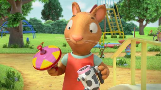 Geschenkt ist geschenkt | Rechte: © Magic Light Pictures LTD 2023 Vor dem Spielplatz betrachtet die Maus Posy erfreut die Spielsachen, die sie in den Händen hält. In ihrer rechten Hand hat sie einen bunten Brummkreisel und in ihrer linken hält sie ein Spielzeug mit Kuhflecken und Haltegriff.
