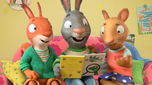 Jamila, das Eichhörnchen, Pip, das Kaninchen und Posy, die Maus, sitzen nebeneinander auf einer pinkfarbenen Couch mit rot-blauen Blumen. Pip hält ein geöffnetes Dino-Buch in beiden Händen.  Alle drei schauen lächelnd in das Buch. Posy hat ihre rechte Hand auf Pips linker Schulter liegen. Ihre linke Hand zeigt geöffnet nach vorne. Neben Jamila liegt ein gelbes Kissen mit grünen Punkten und neben Posy ein grünes Kissen mit Karomuster. An der gelben Wand befinden sich verschiedene Bilder.
