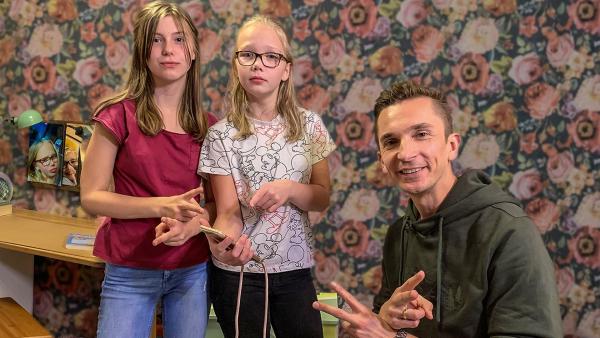 Moderator Eric Mayer mit Julia und Lea vor einer bunten Tapete und einem Schreibtisch im Hintergrund. Lea hält eine Smartphone und zeigt darauf. Julia und Eric machen eine Gebärde.