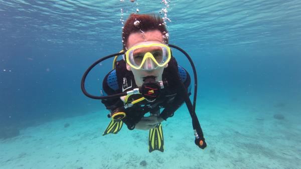 Moderator Eric Mayer taucht in der Karibik und schaut durch seine Taucherbrille in die Kamera. Unter ihm ist der Meeresboden zu sehen.