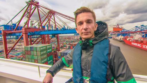 Eric am Hamburger Hafen | Rechte: ZDF Eric Mayer steht angelehnt an ein Geländer. Er blickt den Zuschauer an und blickt von weiter oben, auf einen Teil des Hamburger Hafens. Im Hintergrund sind Schiffe, gestapelte Container und Kräne zu sehen.