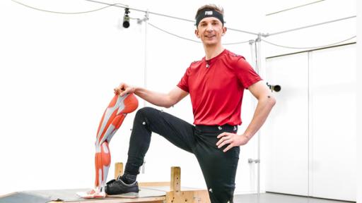 Moderator Eric Mayer steht in Sportkleidung in einem Gymnastikraum. Er hat sein rechtes Bein auf einem Turngerät aufgestützt und hält neben sein Bein das Modell eines künstlichen Beins.