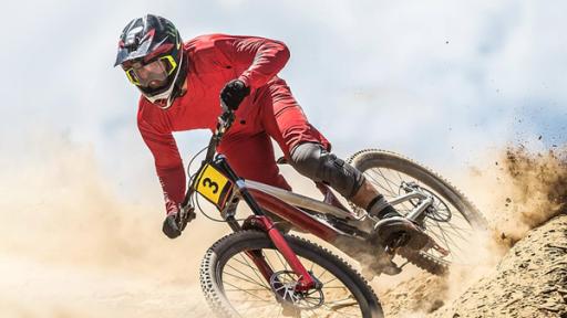 PUR+ - Teaser - Downhill | Rechte: Imago Ein Downhill-Fahrer im roten Anzug und mit Helm fährt auf einem Mountainbike einen Hügel hinab. Er scheint Teilnehmer eines Radrennens zu sein, da sein Fahrrad die Nummer Drei am Lenker trägt. Er wirbelt eine Menge Staub auf.