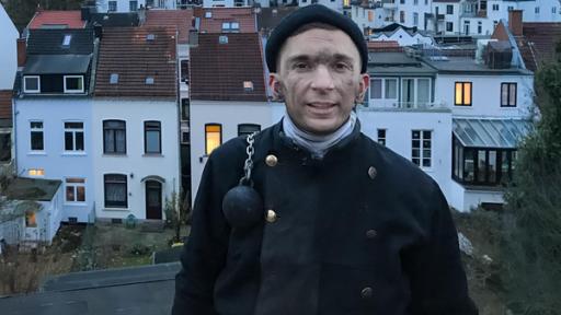 PUR+ - Teaser - Eric wird Schornsteinfeger | Rechte: ZDF PUR+ Modertaor Eric Mayer steht in Schornsteinfeger-Arbeitskleidung und mit rußigem Gesicht in erhöhter Position und schaut in die Kamera. Im Hintergrund sind Häuser zu sehen, bei denen in manchen Fenstern der Schein einer Lampe zu sehen ist.