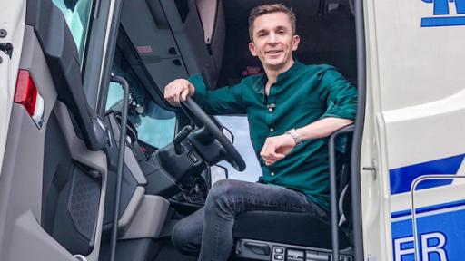 Moderator Eric Mayer sitzt im Fahrerhaus eines weiß-blauen LKW. Die Tür steht offen.