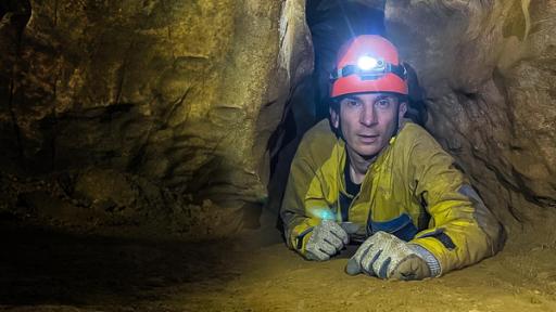 PUR+ - Teaser - Geheimnisvolle Höhlen | Rechte: ZDF Moderator Eric Mayer liegt ausgestattet mit Handschuhen, einem gelben Overall und einem Helm mit Stirnlampe auf dem Boden einer Höhle in einer Höhlenspalte.