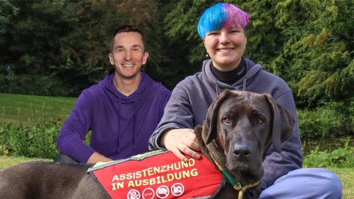 PUR+ - Bühne L - Helfende Hunde -2 | Rechte: ZDF, Collage Assistenzhund Frieda liegt vorne im Bild in der Mitte und trägt ein Geschirr mit der Aufschrift "Assistenzhund in Ausbildung". Dahinter sitzt ihr Frauchen und links daneben Eric. Beide sitzen im Schneidersitz.