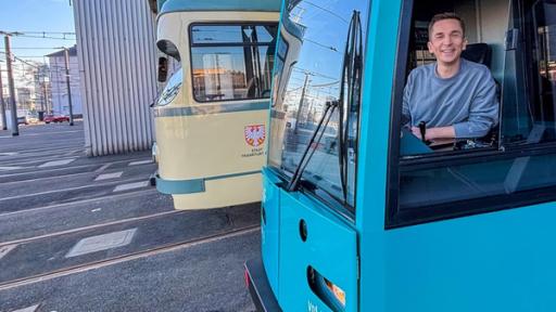 Eric sitzt im Führerhaus einer blauen Straßenbahn. Er schaut durchs offene Fenster in die Kamera und lächelt.
