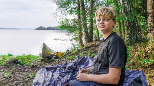 PUR+ - Teaser - Rein ins Abenteuer | Rechte: ZDF Marc sitzt auf einem steinigen Untergrund am Rande eines großen Sees. Neben ihm liegt das noch nicht aufgebaute Zelt. Ein paar Meter weiter liegt ein Kanadier am Ufer. Hinter dem See liegen bewaldete Berge.