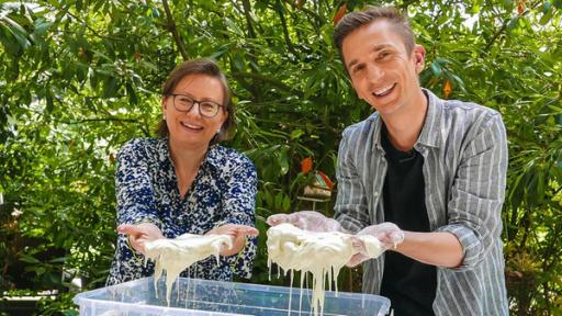 PUR+ - Teaser - Wundermittel Schleim | Rechte: ZDF Eric Mayer (rechts) zusammen mit Wissenschaftsjournalistin Susanne Wedlich (links): Beide stehen an einem Tisch mit einem Becken voll selbst gemachtem Schleim aus Wasser und Maisstärke. Beide halten ihre Hände hoch die voll weißem Schleiß sind und schauen in die Kamera.