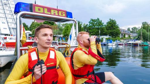Eric sitzt zusammen mit einem Rettungschwimmer der DLRG in einem Boot der DLRG. Der Rettungsschwimmer schaut durchs Fernglas. Eric hät ein Fernglas in der Hand. Beide blicken auf den See hinaus.