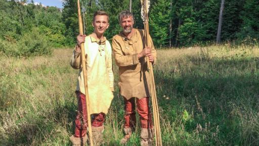 PUR+ - Teaser - Zurück in die Steinzeit | Rechte: ZDF Moderator Eric Mayer steht links im Bild. Er trägt steinzeitiche Kleidung aus Leder und Lederstiefel. In der rechten Hand hält er einen riesigen Speer aus Holz. Rechts nebem ihm steht ebenso gekleidet und mit riesigen Pfeilen in der Hand ein Steinzeitexperte mit ihm auf einer Wiese.