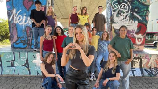 Rechte: KiKA/EBU/Antonia Hirnich Vorne steht Kati K und macht eine Herz-Geste mit ihren Händen. Hinter ihr mehrere Jugendliche gestaffelt in drei Reihen vor Betonpfeilern mit Graffitti. Alle lächeln.