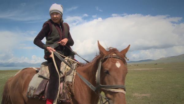 Erkinai sitzt auf ihrem Pferd. Sie trägt eine Mütze und hat die Zügel in der Hand.