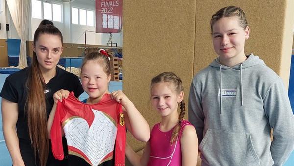 Weltmeisterin Pauline Schäfer-Betz mit Annabelle und ihren Schwestern Estelle und Marielle. Sie stehen in der Trainingshalle. Annabelle hält und präsentiert ihren Nationalmannschaftsanzug.