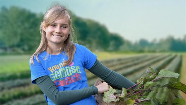 Mira steht lächlend mit einer Kiste Gemüse unter dem Arm auf einem großen Gemüsefeld.