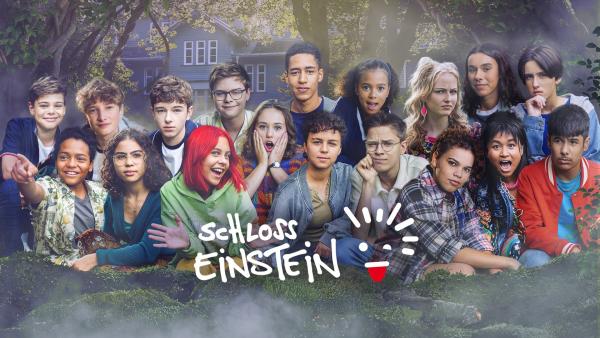 18 Teenager befinden sich eng beieinander auf einem Waldboden. Viele von ihnen lächeln, andere schauen erstaunt oder skeptisch. Um sie herum ist Dunst und hinter ihnen das Schloss-Einstein, umgeben von Bäumen. Unten das Schloss-Einstein-Logo.