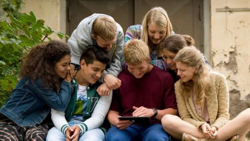 Eine Gruppe junger Menschen sitzt auf einer Bank und schaut lächelnd auf einen Tabletcomputer.
