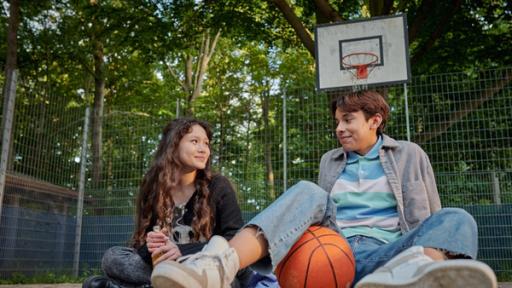 Schloss Einstein (1030) | Rechte: Kika Ein Mädchen und ein Junge sitzen untrer einem Basketballkorb und lächeln sich an.