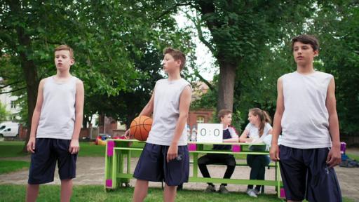 Drei Jungen im Sportdress mit einem Basketball stehen in einem Park. Hinter ihnen ist ein Tisch mit einer Spielstandanzeige aufgebaut.