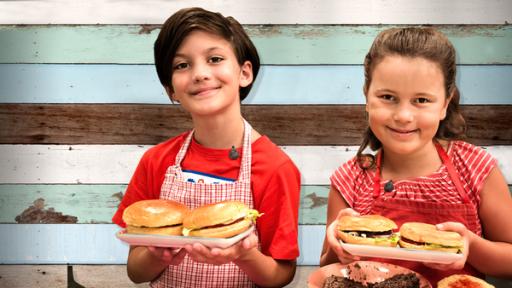Zwei Kinder in roten Schürzen halten Teller mit Burgern und lächeln in die Kamera. 