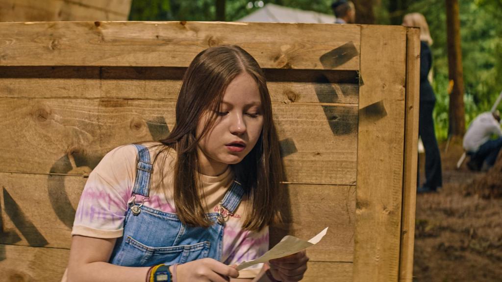 Bei Tageslicht im Wald hockt Meg (Katy Byrne) vor einer großen Holzkiste. Sie schaut auf einen Zettel, den sie in den Händen hält.