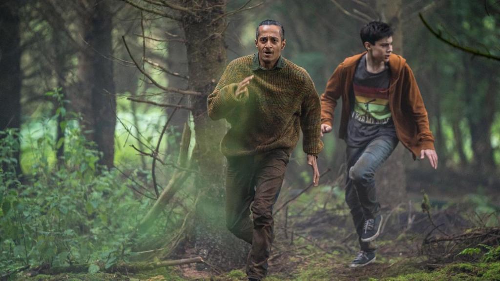 Louis (Oliver Cunliffe) und Fischer (Fayez Bakhsh) laufen bei Tageslicht durch den Wald, der durch einen Grünfilter besonders grün aussieht. Sie sind in Aufruhr mit ängstlich verzerrten Gesichtern.