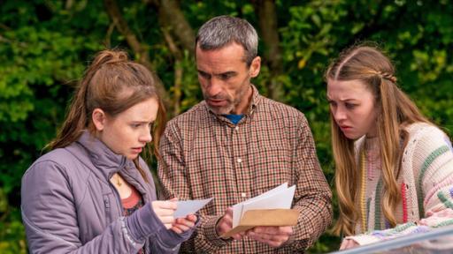 Silverpoint, Staffel 2, Folge 05: Botschaft an mich selbst | Rechte: © Zodiak Kids & Family Productions UK Ltd Draußen neben einem Auto stehen Maeve Vater Aiden, gespielt von Dara Devaney, und Maeve, gespielt von Skye McClenaghan. Auch Bea, dargestellt von Aoife Hughes, steht neben ihnen. Aiden und Bea halten Fotos in den Händen. Aiden schaut zu Bea, die konzentriert auf die Fotos blickt. Maeve sieht von der Seite aus mit angestrengtem Gesicht zu.