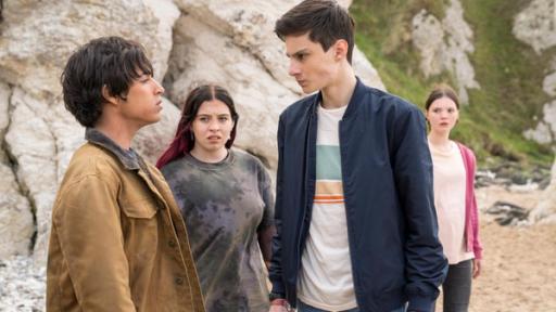 Silverpoint, Staffel 2, Folge 06: Das ist nicht fair! | Rechte: © Zodiak Kids & Family Productions UK Ltd; Stefan Hill Am Strand vor hellem Felsen, stehen sich Louis, gespielt von Oliver Cunliffe, und Jay, gespielt von Noah Manzoor, gegenüber. Louis ist voller Wut mit angespanntem Gesicht und geballten Fäusten. Jay schaut ihn herausfordernd an. Die beiden werden von Kaz, gespielt von Maiya Silveston, und Isabel, gespielt vo Alis Waters, beobachtet. Kaz schaut mit einem verächtlichen Blick zu Jay.