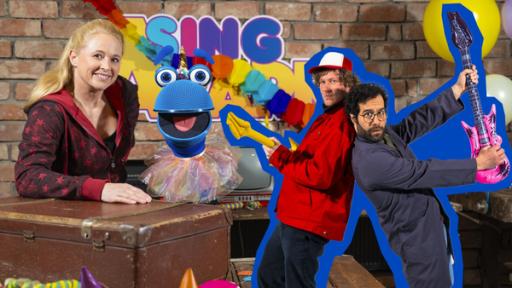Singa und das SingDing schauen lachend hinter einem bunt dekorierten Tisch hervor. Das SingDing trägt ein buntes Tutu. Rechts von den beiden stehen Deniz & Ove, die mit aufblasbaren Gitarren posieren.