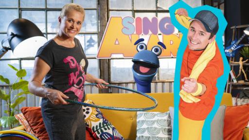 Sing! Hüpf! Tanz! | Rechte: ZDF Singa steht vor einem gelben Sofa und hält mit beiden Händen einen Hula-Hoop-Reifen um ihrer Hüfte fest. Sie trägt ein schwarzes T-Shirt und lächelt mit leicht geneigtem Kopf in die Kamera. Donikkl steht neben ihr und lächelt ebenfalls in die Kamera. Er trägt ein oranges T-Shirt, eine blaue Kappe und hält mit den Händen ein gelbes Handtuch über seinen Schultern fest. Das SingDing schaut zwischen den beiden hindurch hinter dem Sofa hervor und blickt mit offenem Mund in die Kamera.