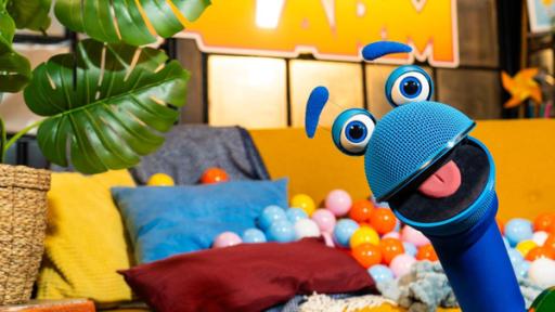 SingDings Spezial Sommersehnsucht | Rechte: ZDF Das SingDing schaut mit weit geöffnetem Mund in die Kamera. Im Hintergrund steht ein Sofa, auf dem mehrere Kissen und viele bunte Plastikbälle liegen. Links im Bild steht eine grüne Pflanze.